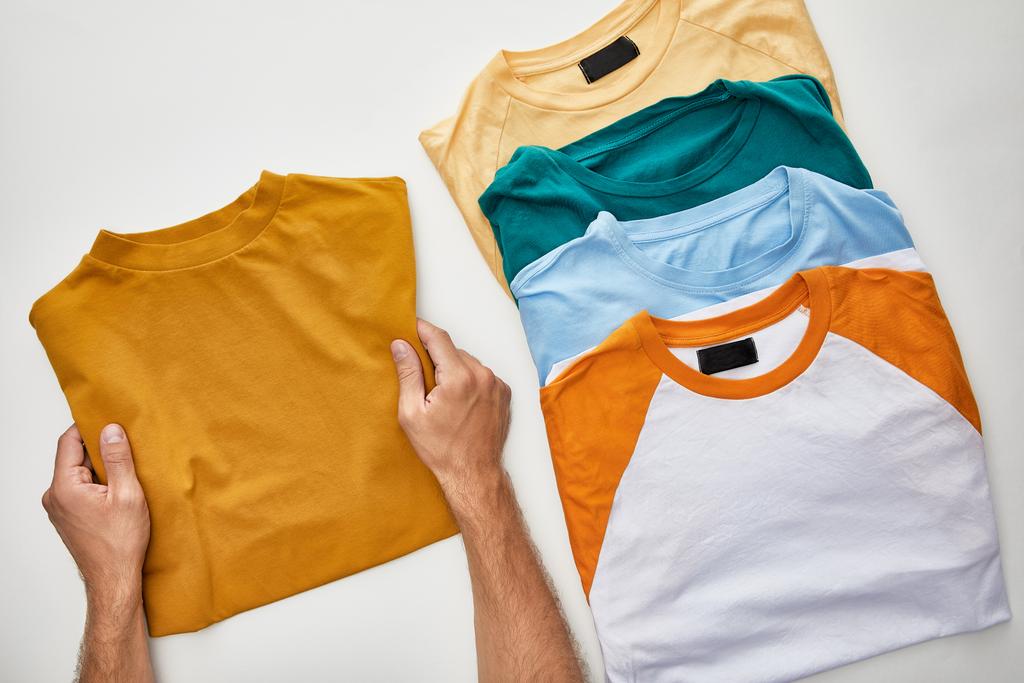 cropped view of man holding ochre t-short near beige, orange, turquoise and blue ones on white background - Photo, Image
