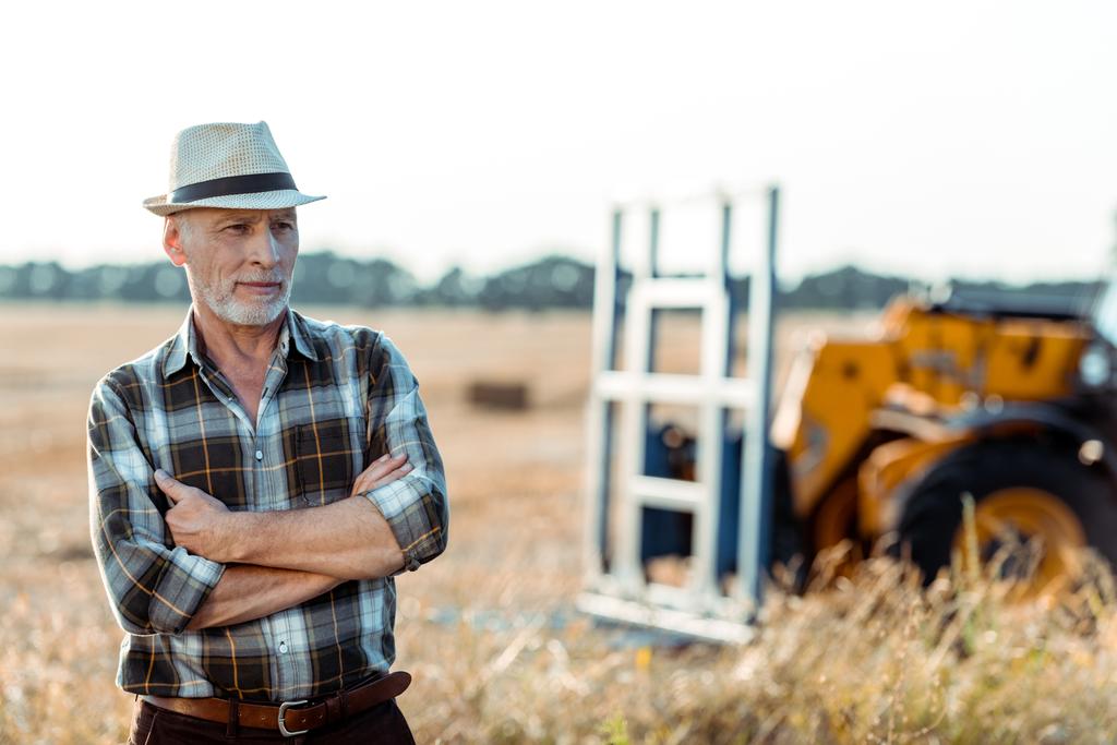самозанятый фермер в соломенной шляпе стоит со скрещенными руками возле трактора
  - Фото, изображение