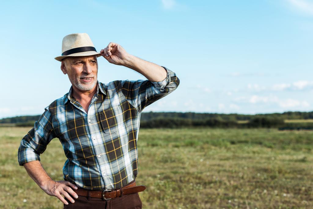 χαρούμενος αγρότης στέκεται με το χέρι στο ισχίο, ενώ αγγίζοντας ψάθινο καπέλο  - Φωτογραφία, εικόνα