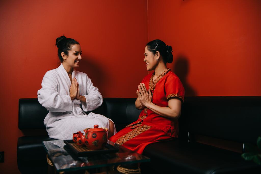 笑顔の女性とアジアのマッサージ師がソファに座って挨拶の手を見せる - 写真・画像