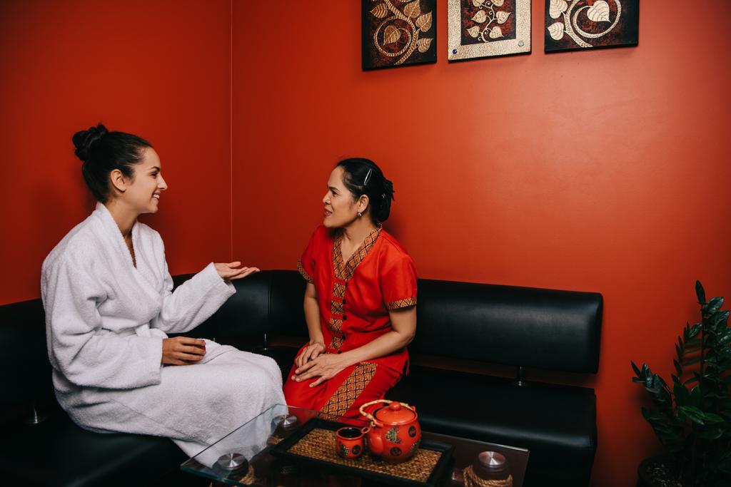 lächelnde Frau und asiatischer Masseur sitzen auf dem Sofa und unterhalten sich im Wellnessbereich  - Foto, Bild