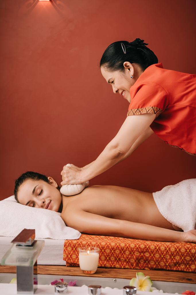 Zijaanzicht van Aziatische masseur doet terug massage met kruiden ballen naar vrouw in Spa - Foto, afbeelding