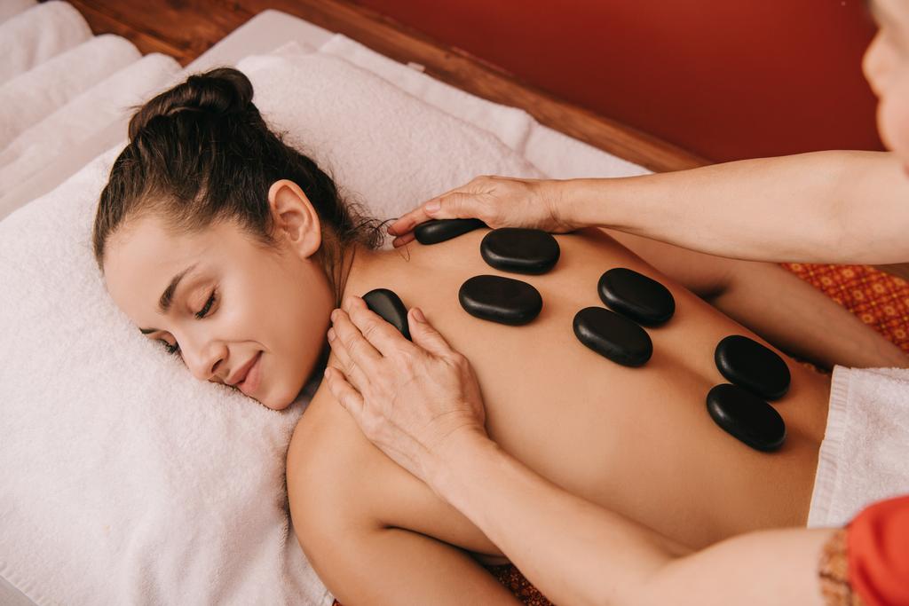 vista cortada de massagista fazendo massagem de pedra quente para a mulher no salão de spa
  - Foto, Imagem