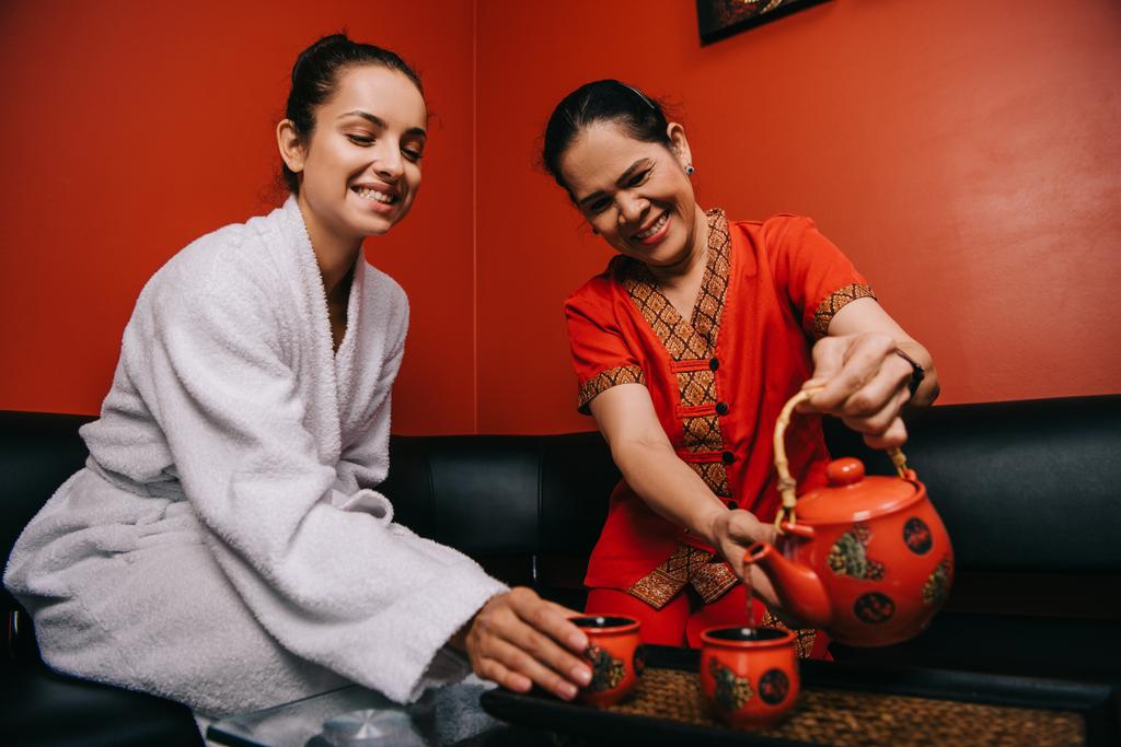 азіатський масажист наливає чай на чашку і приваблива жінка в халаті посміхається в спа-центрі
  - Фото, зображення