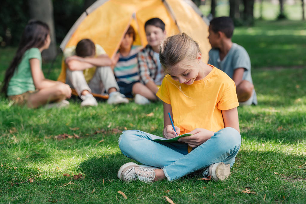 Selektiver Fokus glücklicher Kinder, die in der Nähe multikultureller Freunde im Camp in Notizbücher schreiben  - Foto, Bild