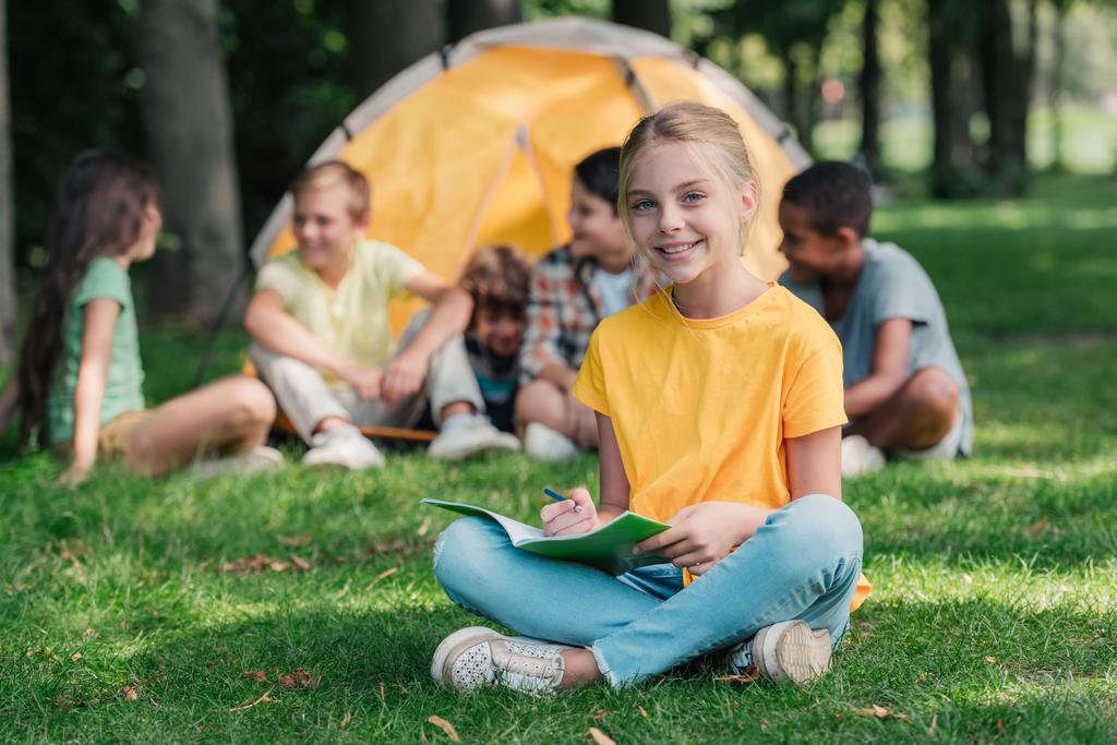 selective focus of cheerful kid holding notebook near multicultural friends in camp  - Photo, Image