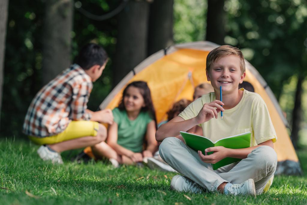 foco seletivo de criança feliz segurando notebook e lápis perto de amigos no acampamento
  - Foto, Imagem