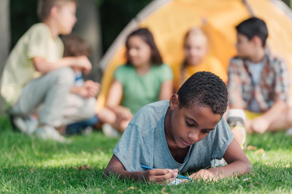 foyer sélectif de heureux afro-américain garçon écrit dans un carnet près des amis dans le camp
  - Photo, image