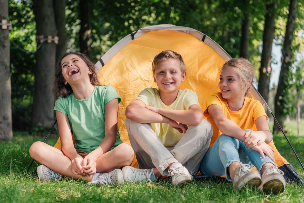 счастливый мальчик, сидящий с симпатичными друзьями, смеющийся возле лагеря
  - Фото, изображение