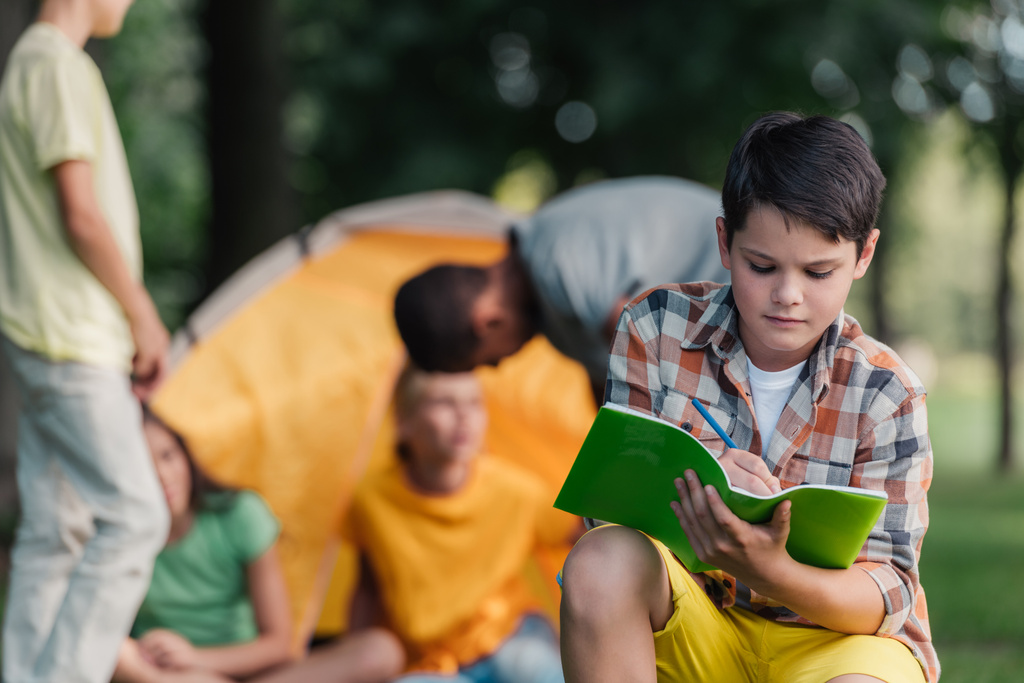 enfoque selectivo de niño sosteniendo cuaderno y lápiz cerca de amigos en el campamento
  - Foto, imagen