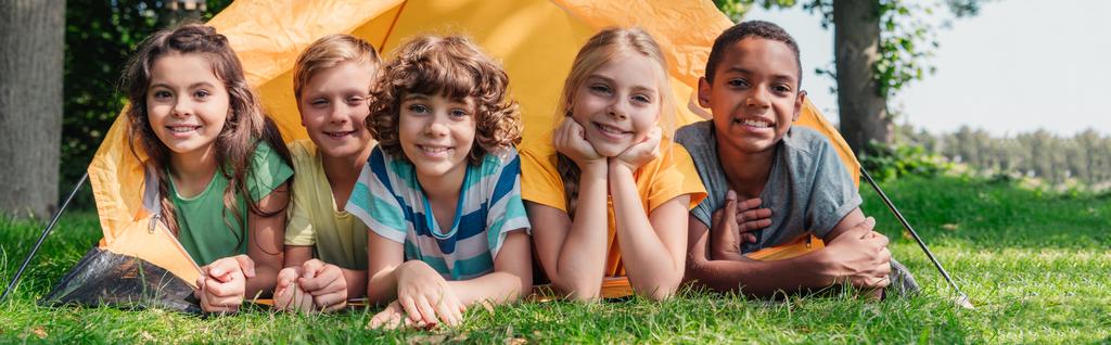 tiro panorámico de niños felices multiculturales sonriendo mientras yacen cerca del campamento
  - Foto, Imagen