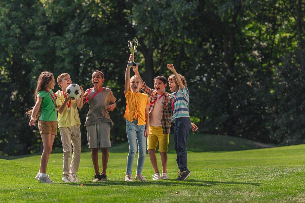 ευτυχισμένα πολυπολιτισμικά παιδιά στέκονται στο γρασίδι με το ποδόσφαιρο και τρόπαιο  - Φωτογραφία, εικόνα