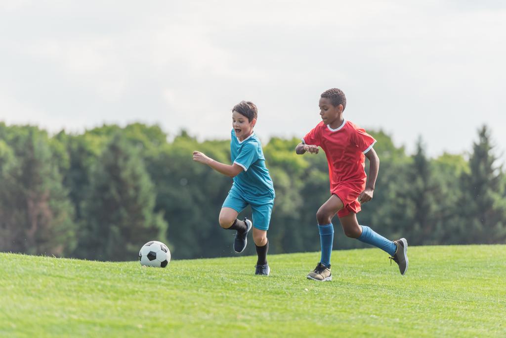 vzrušené multikulturní děti hrající fotbal na trávě  - Fotografie, Obrázek