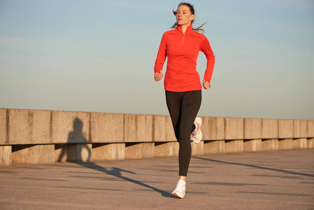 Uma mulher de corrida de camisa vermelha e pernas pretas na rua ao nascer do sol. Correndo em cais de concreto
 - Foto, Imagem