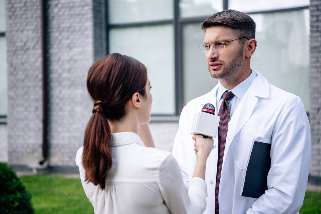 журналист держит микрофон и разговаривает с красивым доктором в белом халате
 - Фото, изображение