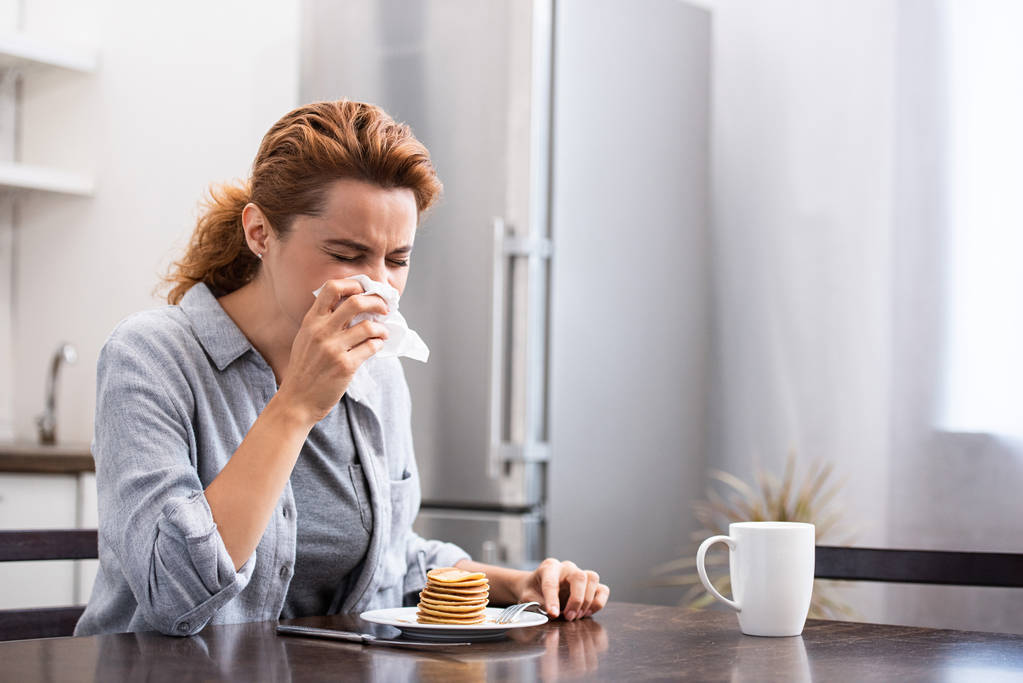 sick woman sneezing in tissue near sweet pancakes at home  - Photo, Image