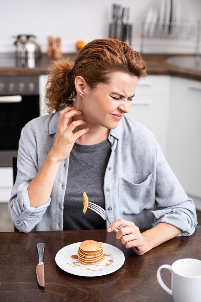 Frau mit Allergie kratzt Hals, während sie Gabel in der Nähe von Pfannkuchen hält  - Foto, Bild