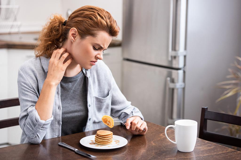 attraktive Frau mit Allergie kratzt Hals, während sie Gabel mit Pfannkuchen hält  - Foto, Bild
