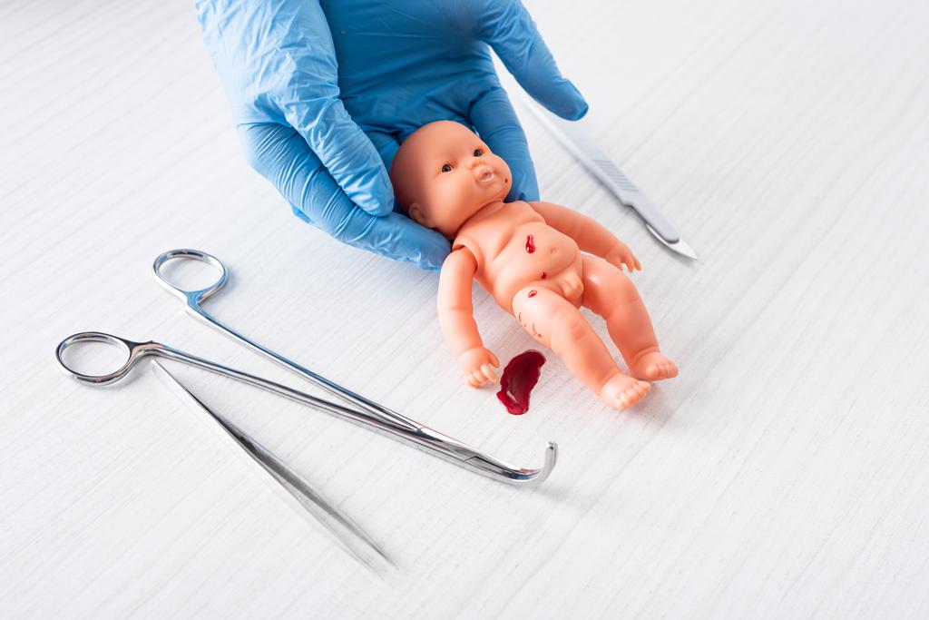 vista ritagliata del medico in guanti di lattice blu in possesso di bambola bambino con sangue vicino a strumenti medici
  - Foto, immagini