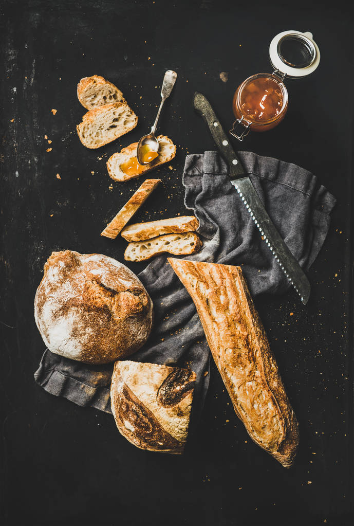 Επίπεδη όψη φρεσκοψημένου μπαγκέτα σε φέτες, ρολό ψωμιού και μαρμελάδα βερίκοκο σε σκούρο λινό πάνω από ρουστίκ μαύρο φόντο, κορυφή θέα - Φωτογραφία, εικόνα