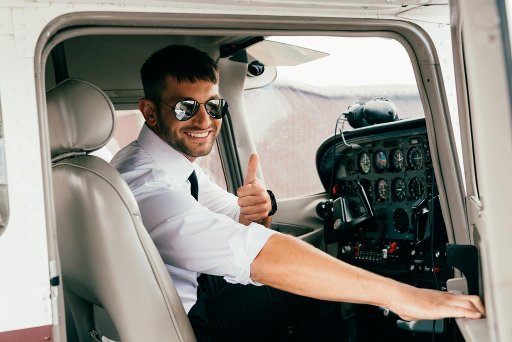 smiling handsome pilot in sunglasses looking at camera and showing thumb up in plane - Photo, Image