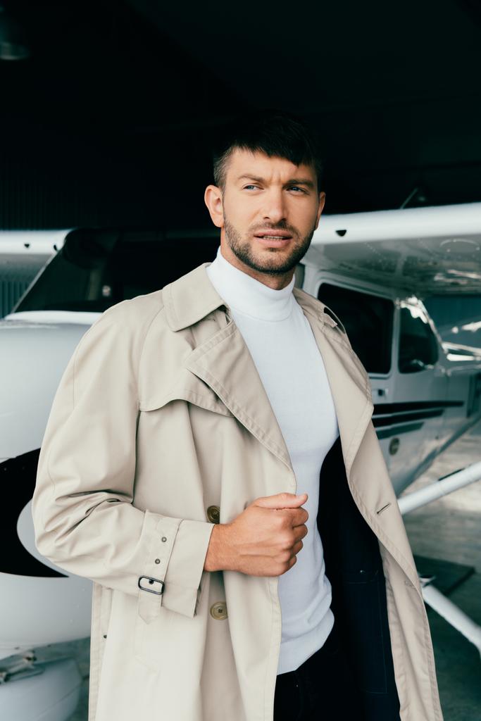 peinzende stijlvolle zakenman in vacht staande in de buurt van vliegtuig en op zoek weg - Foto, afbeelding