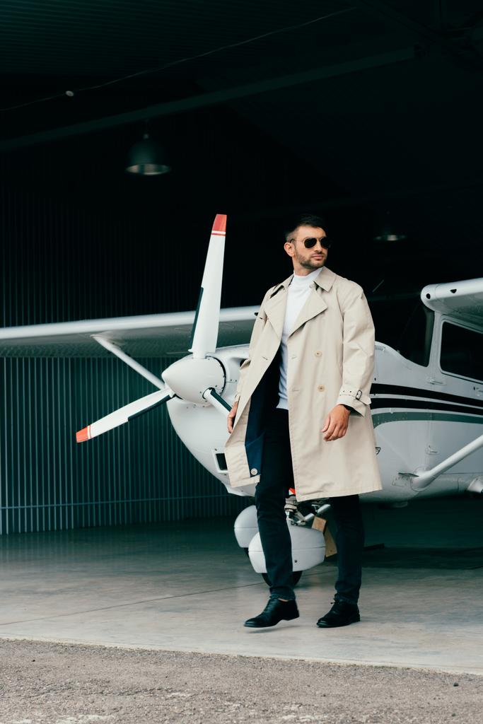 Ganzkörperansicht des stilvollen Geschäftsmannes im Mantel, der in der Nähe von Flugzeug steht - Foto, Bild