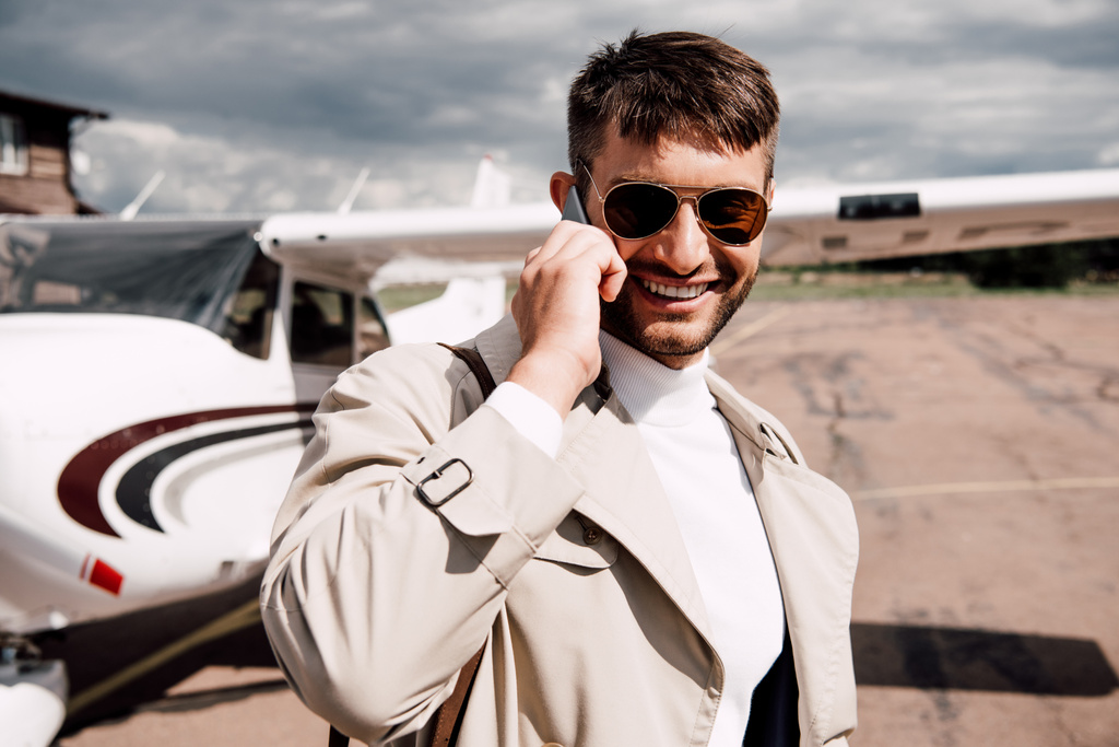 Χαμογελαστός άνθρωπος με παλτό με τσάντα που μιλάει στο smartphone κοντά στο αεροπλάνο - Φωτογραφία, εικόνα