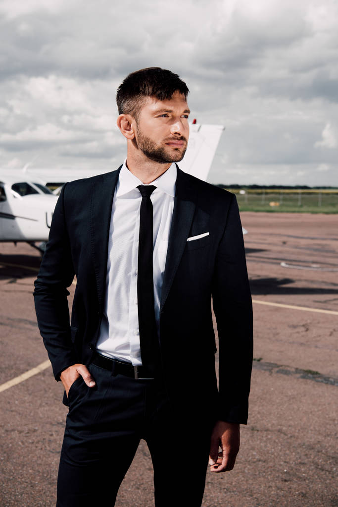 Selbstbewusster bärtiger Geschäftsmann in formeller Kleidung, der mit der Hand in der Tasche in der Nähe des Flugzeugs steht - Foto, Bild