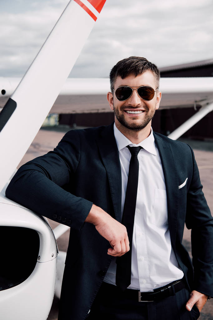 Улыбающийся бизнесмен в формальной одежде стоит с рукой в кармане рядом с самолетом
 - Фото, изображение