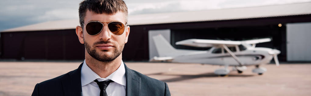 panoramische foto van serieuze zakenman in formele slijtage en zonnebrillen staande in de buurt van het vliegtuig - Foto, afbeelding
