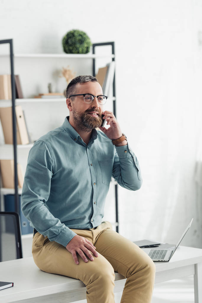 handsome businessman in shirt and glasses sitting on table and talking on smartphone - Zdjęcie, obraz