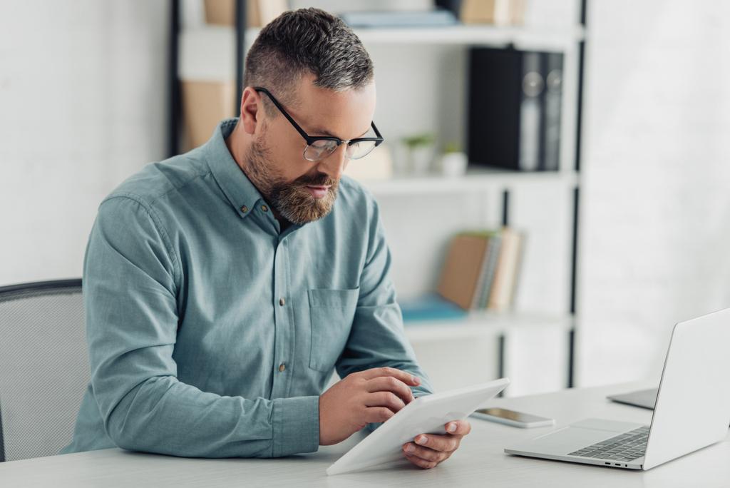 όμορφος επιχειρηματίας σε πουκάμισο και γυαλιά χρησιμοποιώντας ψηφιακό tablet στο γραφείο  - Φωτογραφία, εικόνα