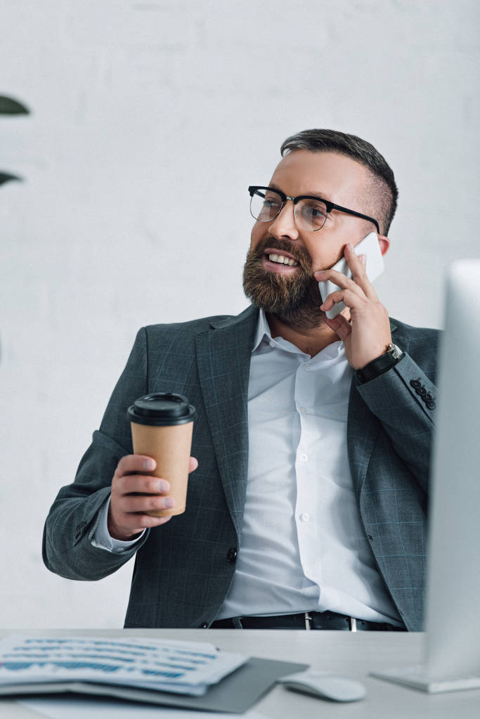 guapo hombre de negocios en ropa formal y gafas hablando en el teléfono inteligente y sosteniendo la taza de papel
 - Foto, Imagen