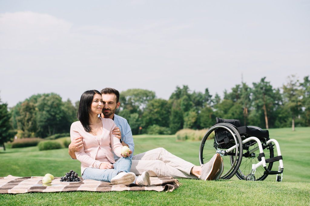 feliz guapo hombre abrazando joven discapacitado novia mientras sentado en manta en parque
 - Foto, imagen