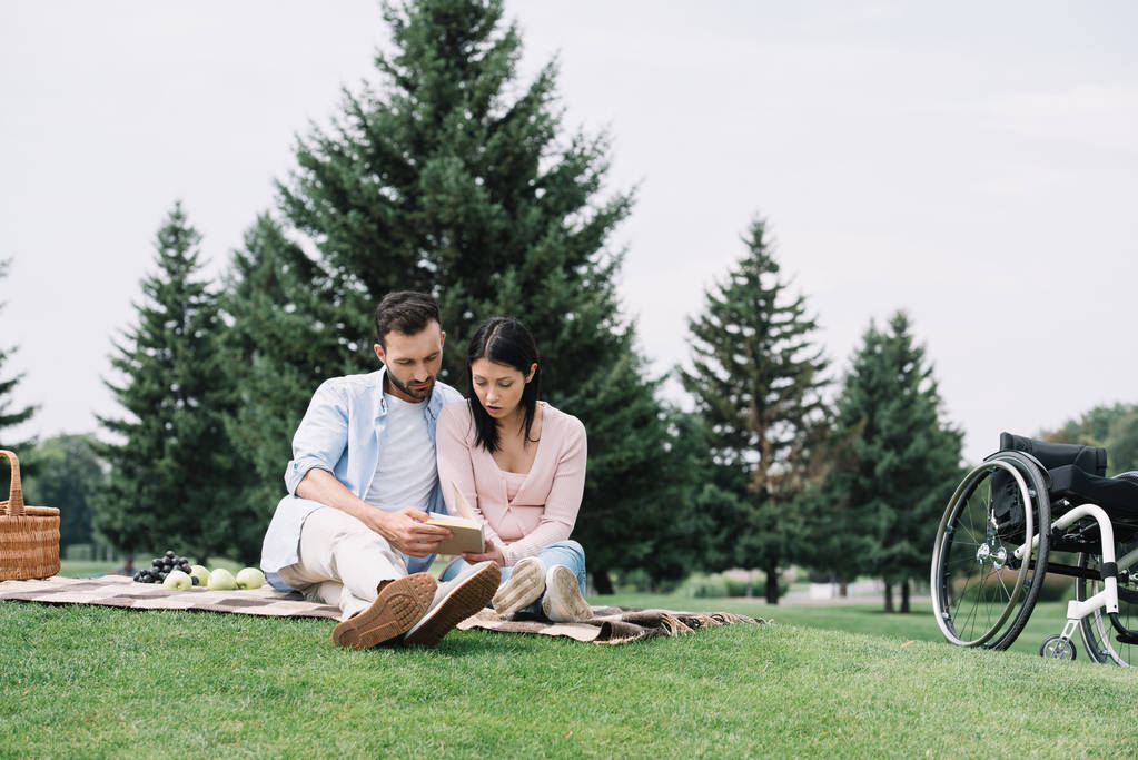 handsome man with disabled girlfriend reading book while sitting on blanket in park - Photo, Image