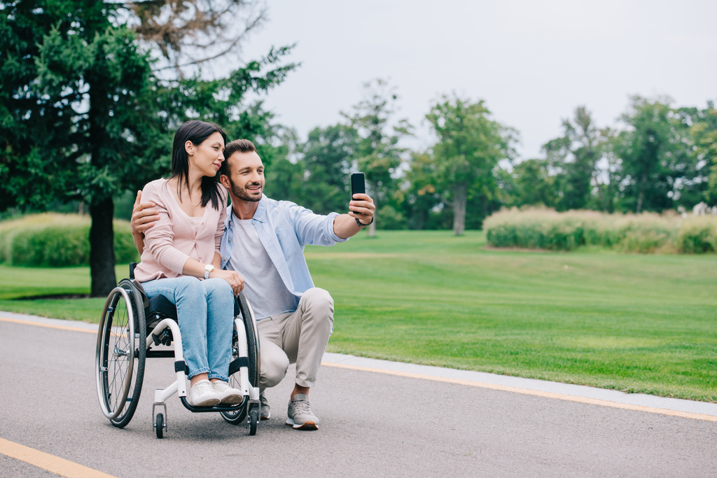 gelukkige man knuffelen gehandicapte vriendin en het nemen van selfie op smartphone in Park - Foto, afbeelding
