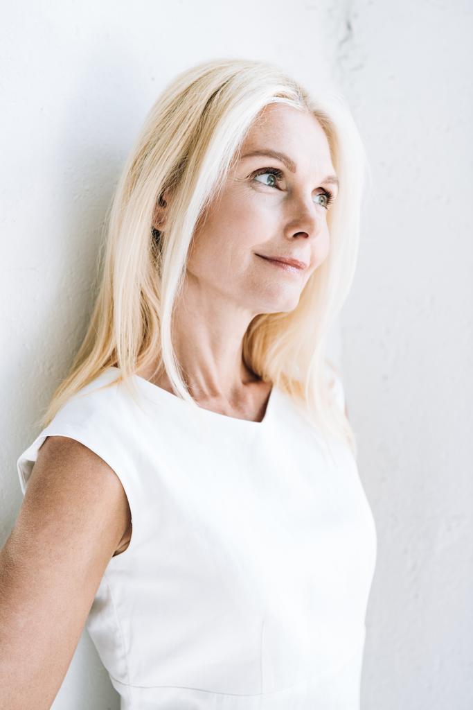 smiling blonde mature woman looking away near white wall - Photo, Image
