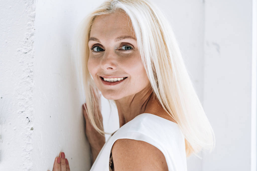onnellinen blondi kypsä nainen poseeraa lähellä valkoista seinää
 - Valokuva, kuva