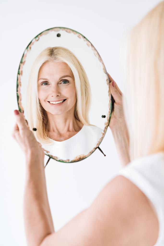 χαρούμενη ξανθιά ώριμη γυναίκα κοιτάζοντας μέσα καθρέφτη απομονώνεται σε λευκό - Φωτογραφία, εικόνα