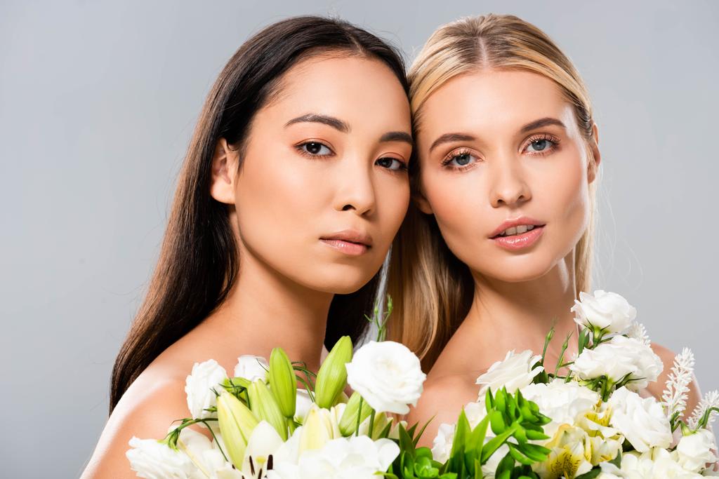 mooie blonde en Aziatische brunette naakte vrouwen met witte bloemen geïsoleerd op grijs - Foto, afbeelding