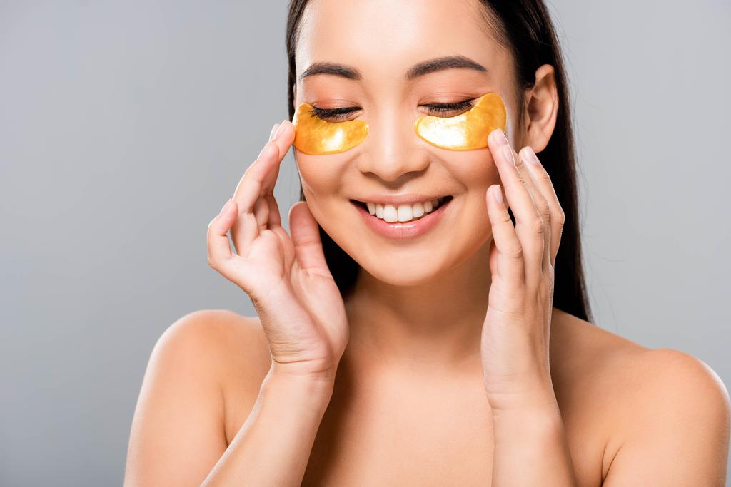 glimlachend naakte Aziatische vrouw met oog patches op gezicht geïsoleerd op grijs - Foto, afbeelding