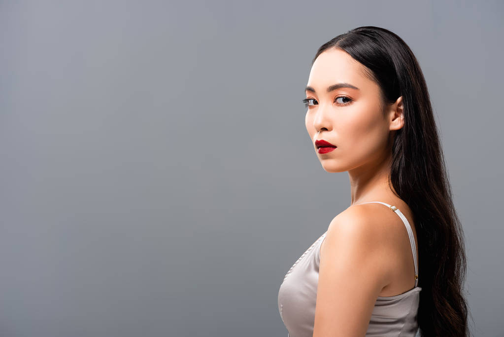 schöne asiatische Frau im Satinkleid mit roten Lippen, die in die Kamera schaut, isoliert auf grau - Foto, Bild