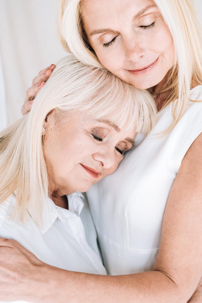 blonde reife Tochter und ältere Mutter umarmen sich mit geschlossenen Augen - Foto, Bild