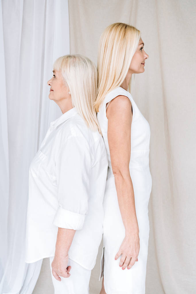 vista laterale della figlia matura bionda e la madre anziana in abiti bianchi totali in piedi schiena a schiena
 - Foto, immagini