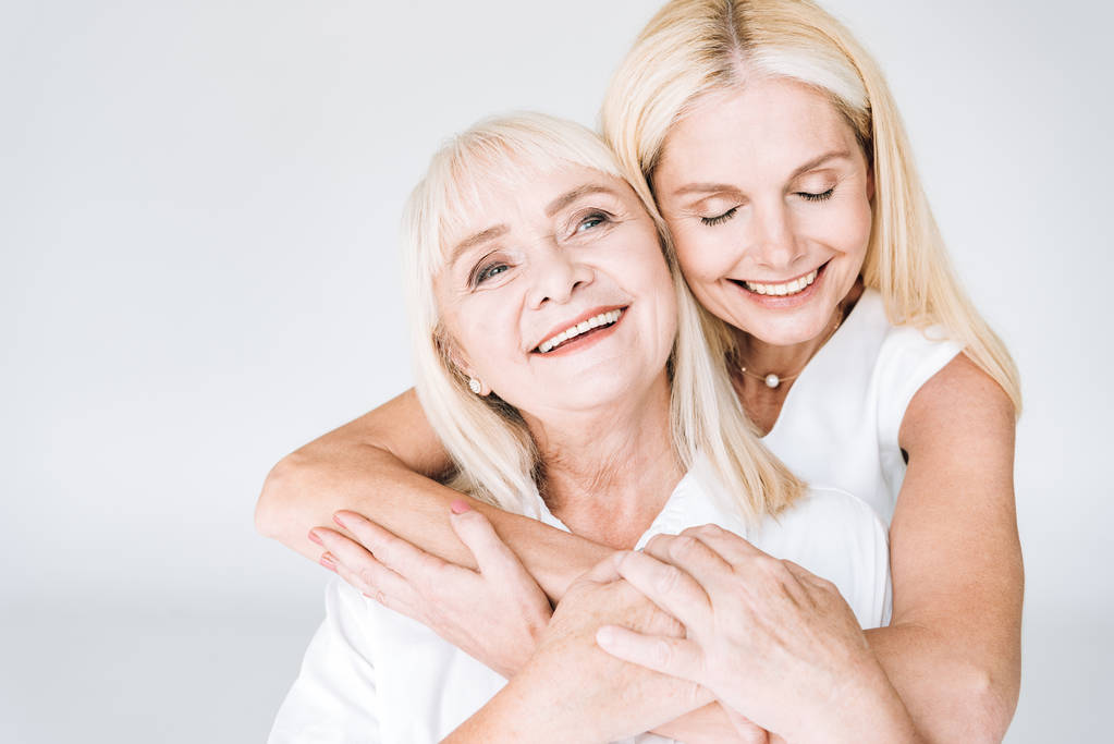 Gelukkig blonde volwassen dochter en senior moeder in totaal witte outfits omarmen geïsoleerd op grijs - Foto, afbeelding