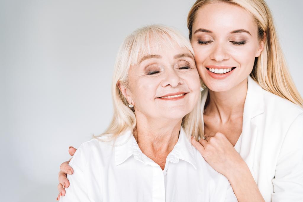 gelukkige blonde grootmoeder en kleindochter samen in totaal witte outfits met gesloten ogen omarmen geïsoleerd op grijs - Foto, afbeelding