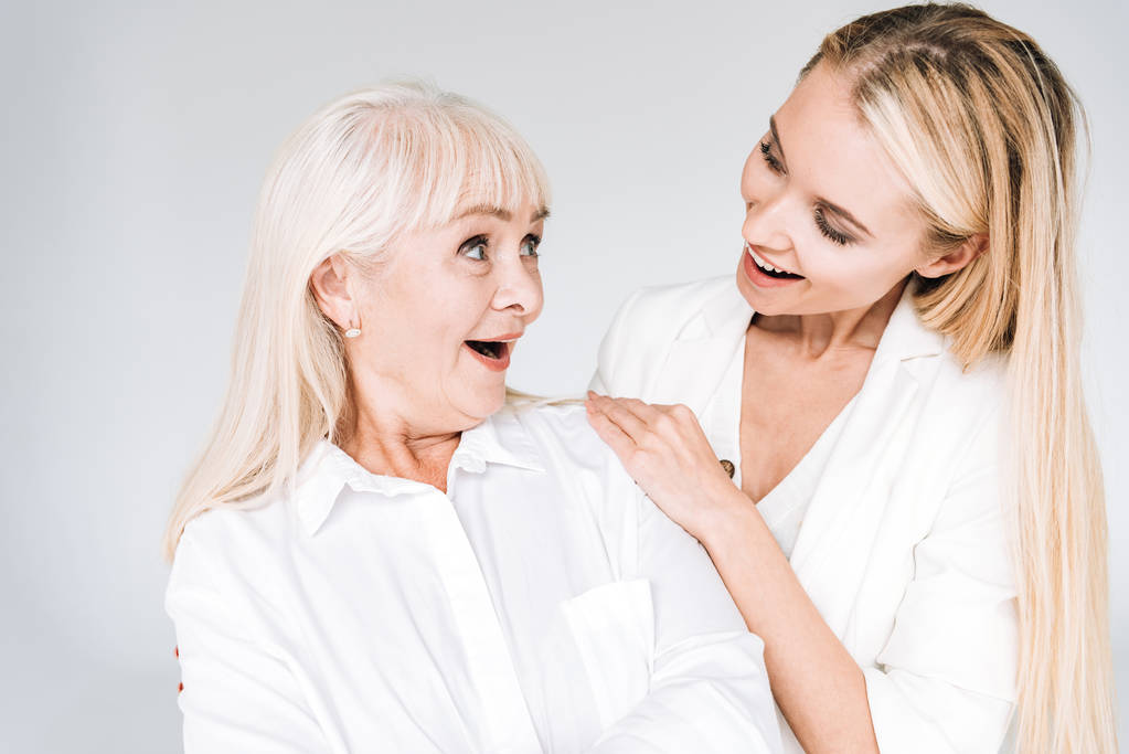 excited blonde grandmother and granddaughter together in total white outfits looking at each other isolated on grey - Photo, Image