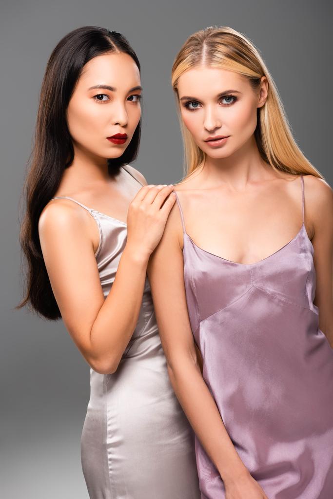 elegante europäische blonde und asiatische brünette Frauen in Satinkleidern isoliert auf grau - Foto, Bild