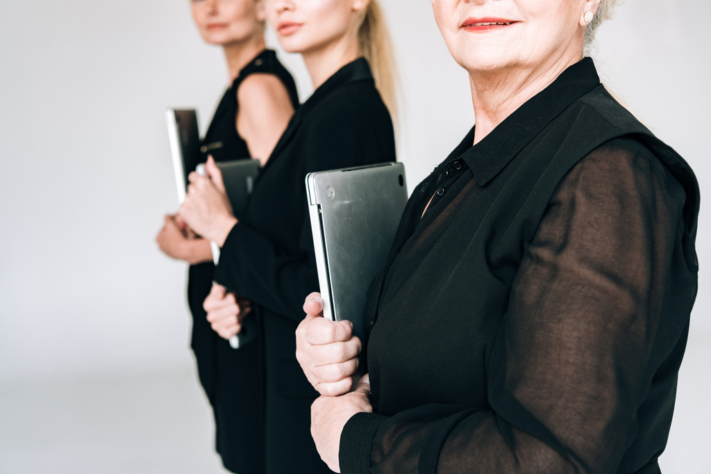 обрізаний вид блондинки трьох поколінь в загальному чорному вбранні, що тримає ноутбуки ізольовані на сірому
 - Фото, зображення